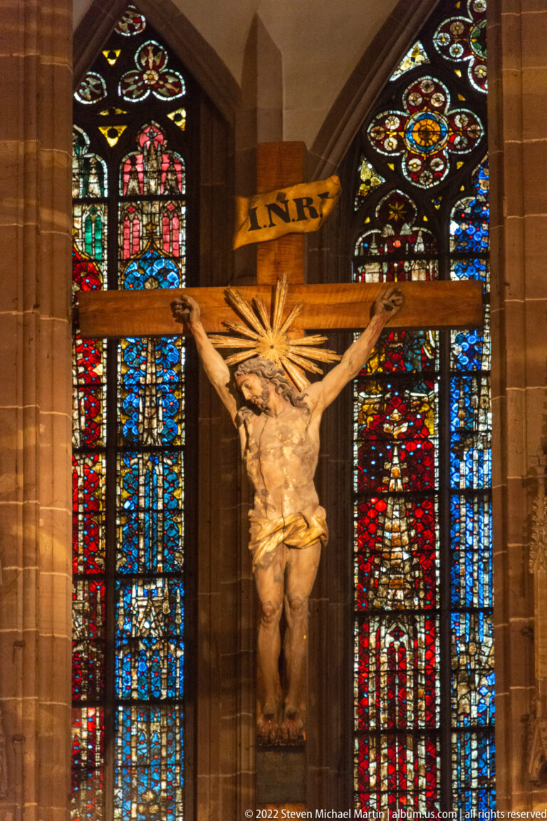 Crucified Christ on a wooden cross stands before brightly colored stained-glass windows in Strasbourg Cathedral.