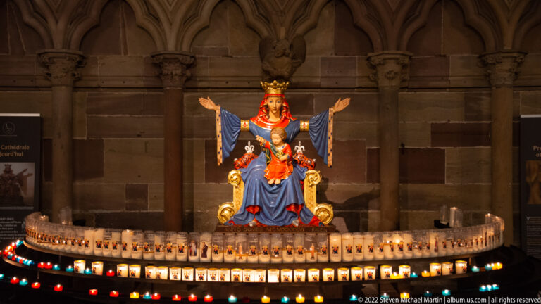Crowned Virgin and Child statue in blue and red robes above rows of glowing votive candles.