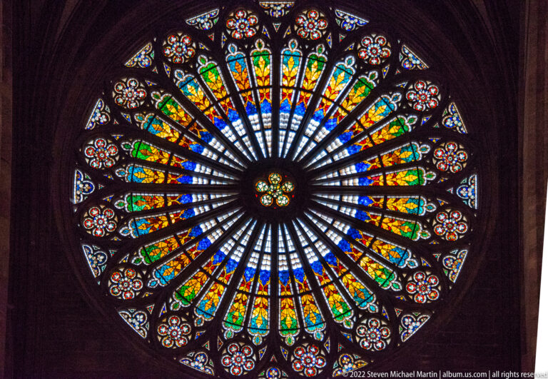 Close frontal view of a circular stained-glass rose window with radiating stone tracery and multicolored panels.