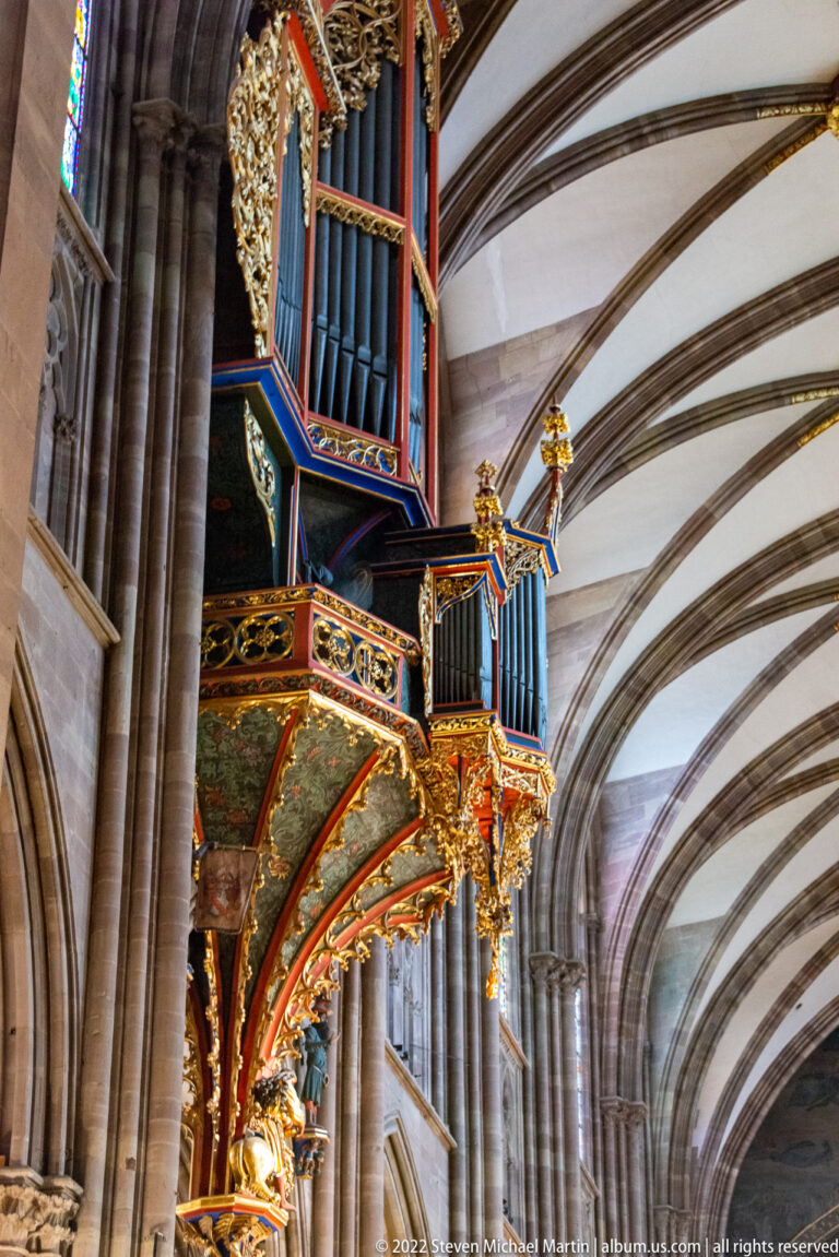 Colorful suspended pipe organ with blue pipes and gilded carved case projecting into the cathedral nave.