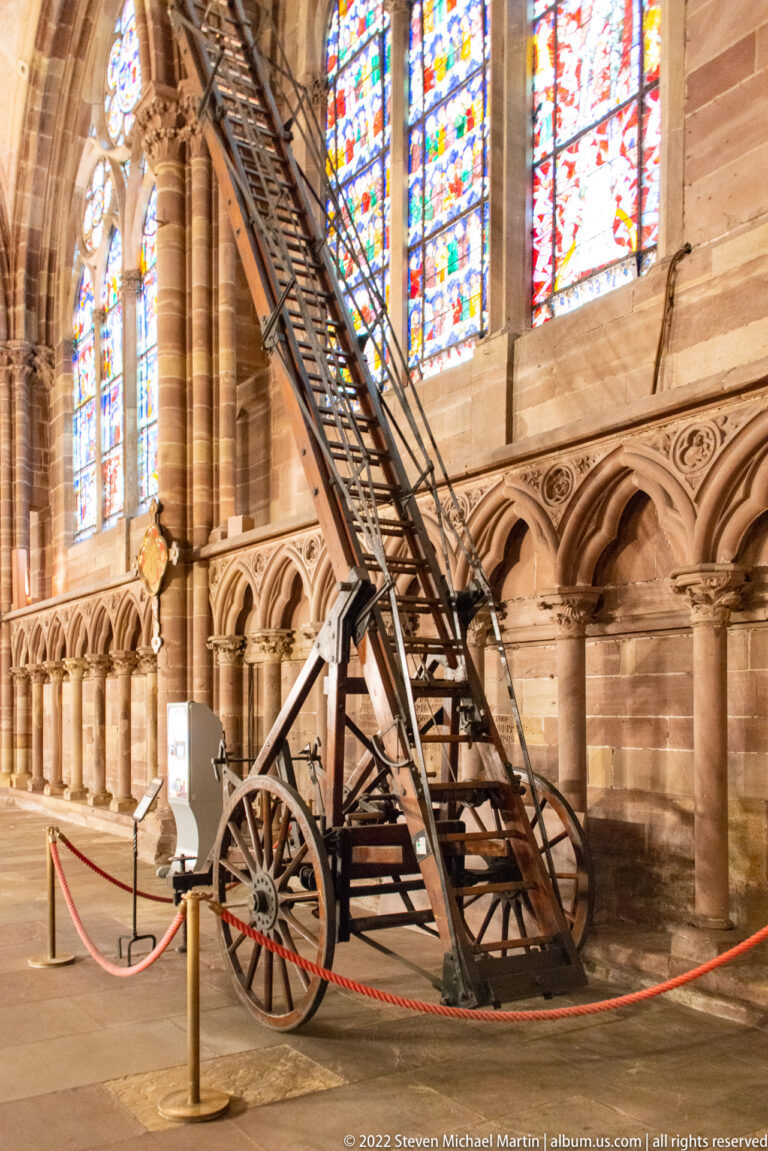 Long wooden ladder on a wheeled carriage displayed beneath stained-glass windows inside Strasbourg Cathedral.