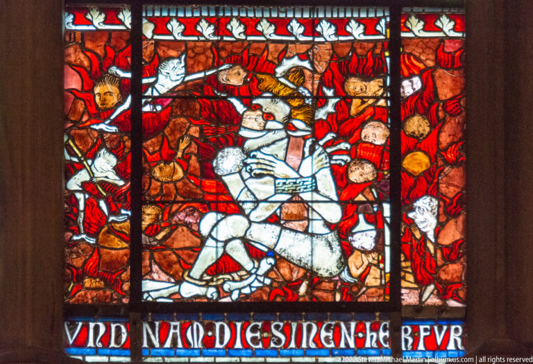 Stained-glass panel with an armored rider on a pale horse amid red figures and a Gothic-script inscription.