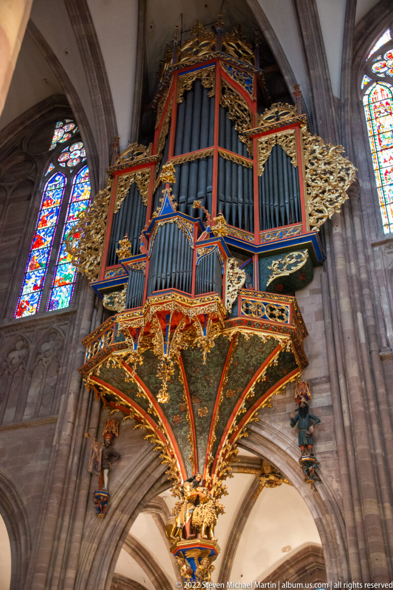 Low-angle view of the cathedral's colorful suspended pipe organ, showing blue pipes, gilded carving, and its ornate underside.