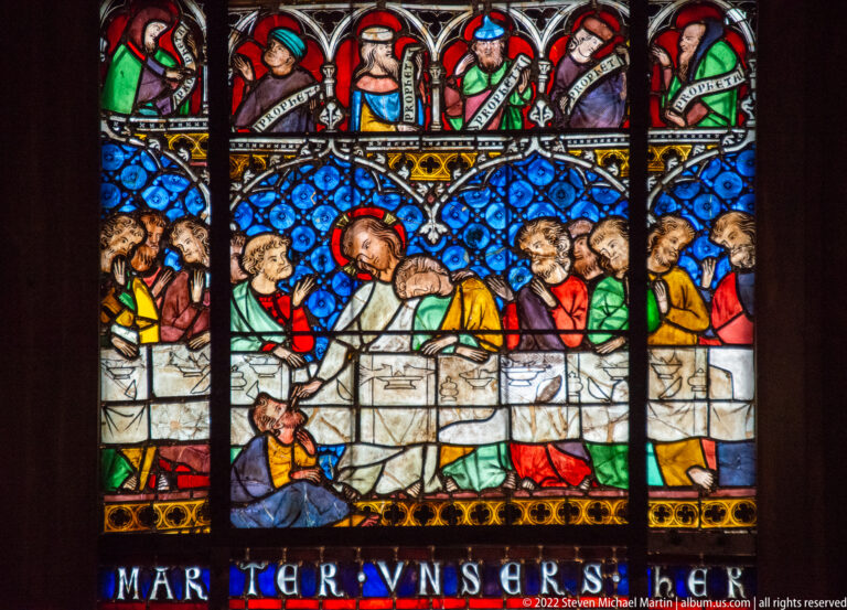 Stained-glass scene of a central sacred figure speaking among clustered men, with portrait figures above and inscription below.