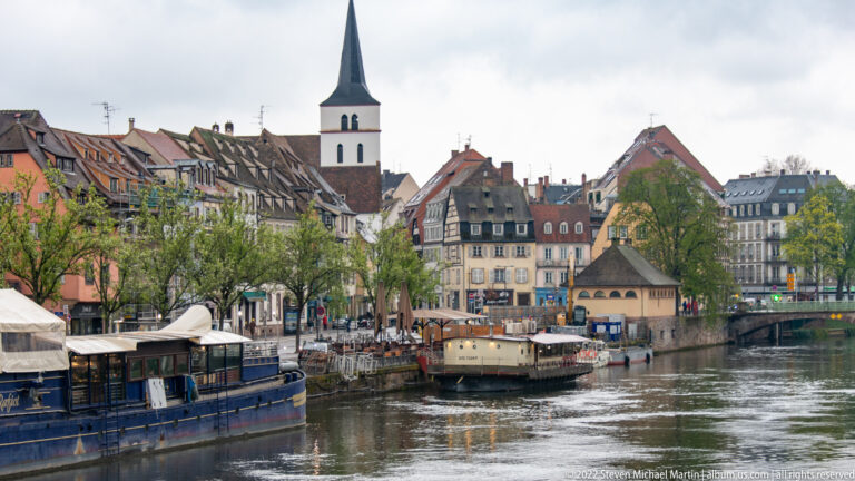 Strasbourg canal bordered by historic buildings, moored boats, trees, and a slender church spire.
