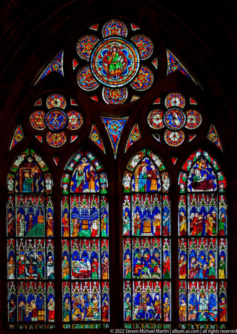 Four tall stained-glass lancets beneath circular and multifoil windows in the dark cathedral interior.
