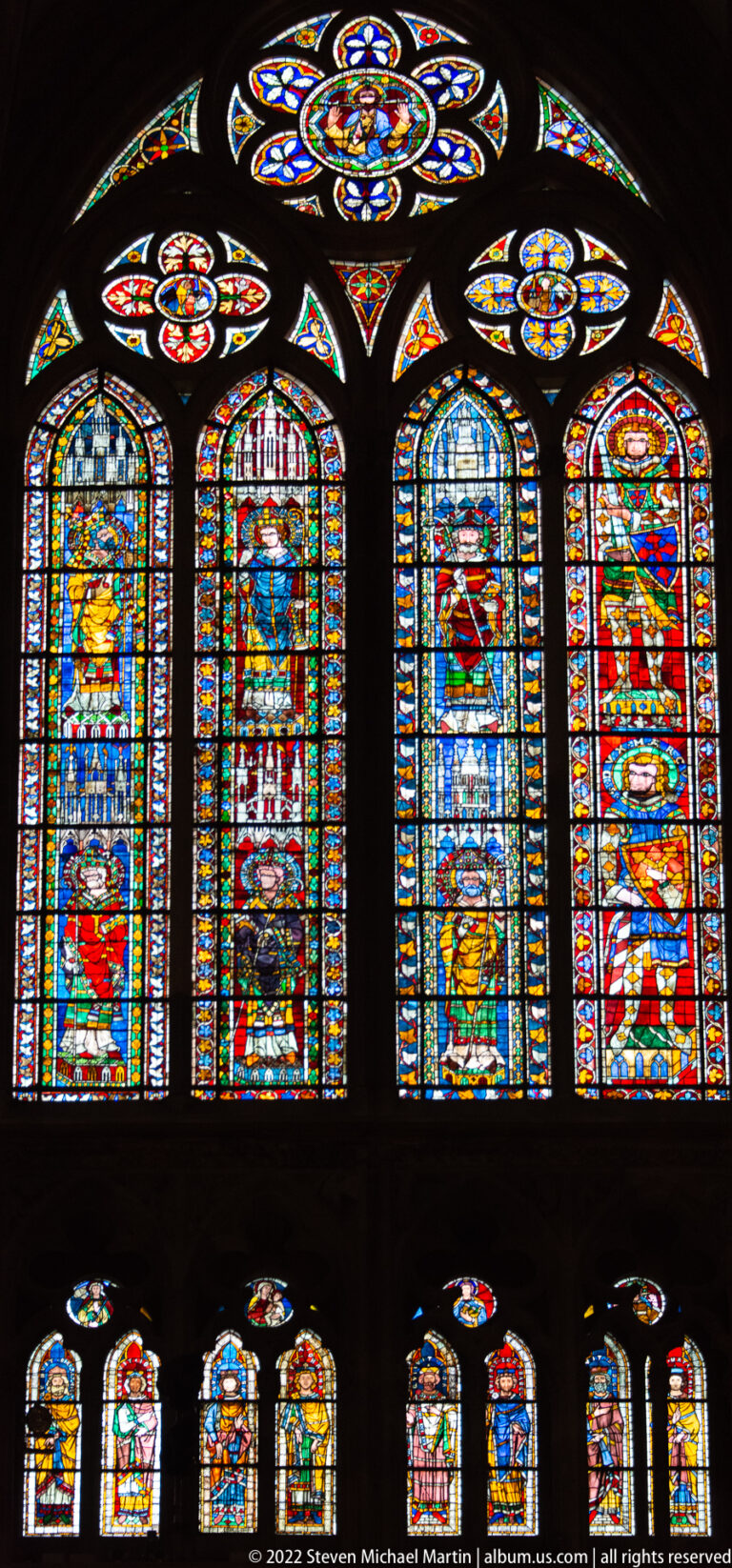 Wide interior view of tall narrative stained-glass lancets with circular tracery lights and smaller windows below.