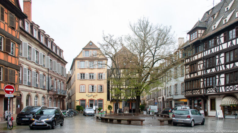 Narrow Strasbourg street lined with half-timbered and plastered houses, parked cars, bicycles, and leafless trees.