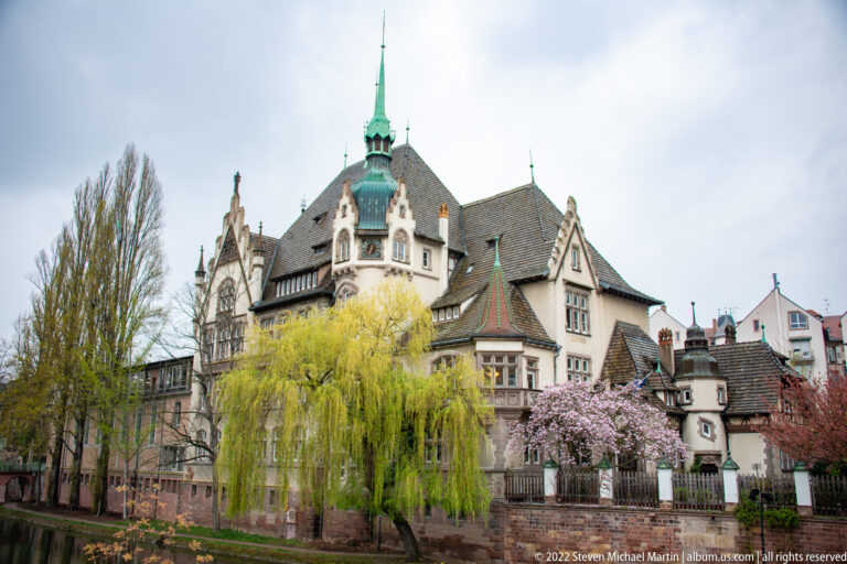 Historic steep-roofed building with decorative gables and green turret behind willow and flowering trees.