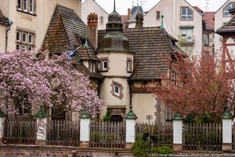 Historic stucco houses with steep tiled roofs and a small round tower framed by pink spring blossoms.