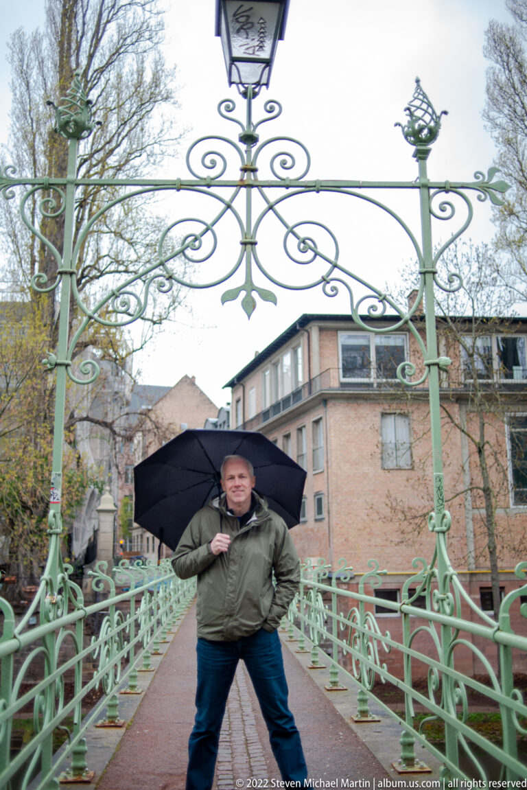 Man holding a black umbrella stands at the center of an ornate green metal footbridge on a rainy day.