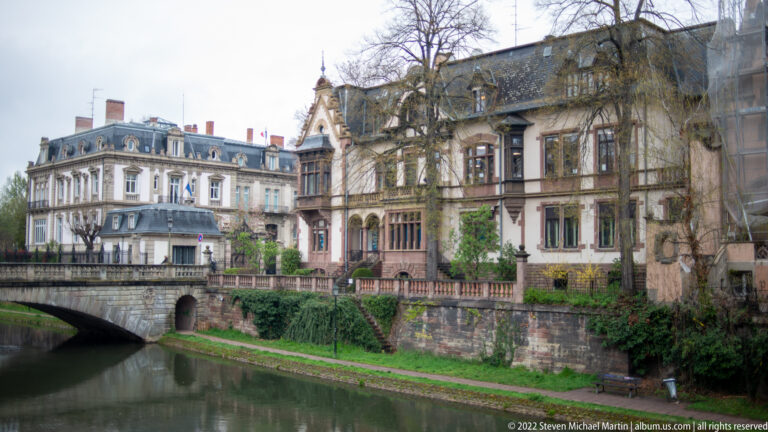 Historic multi-story buildings and a low bridge line a narrow Strasbourg canal beneath bare trees.