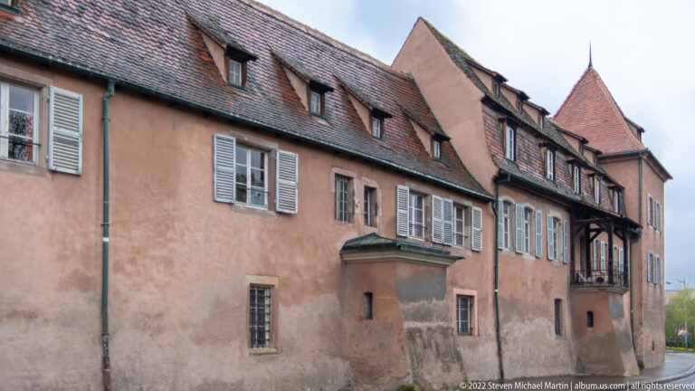 Long salmon-colored historic building with shuttered windows, steep tiled roof, dormers, and a projecting corner tower.