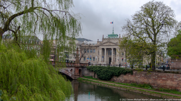 Columned civic building with a French flag seen across a canal, framed by a willow tree and stone embankment.