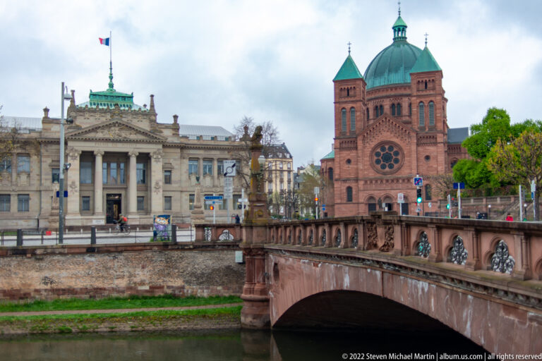 Stone bridge over a canal with a red-sandstone twin-towered church and large civic building beyond.