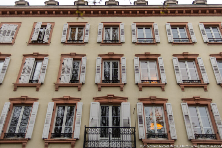 Historic pale stucco façade with rows of shuttered windows, red stone trim, and wrought-iron railings.