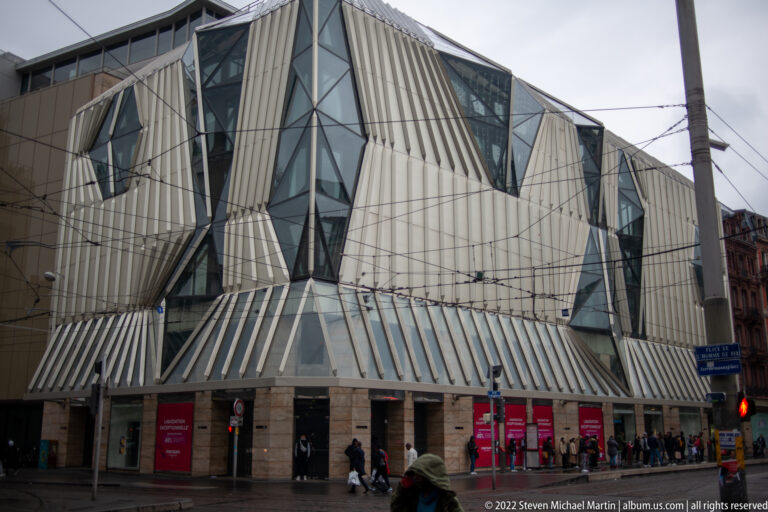 Angular modern building with pale metal cladding and triangular glass bays beneath overhead tram wires.