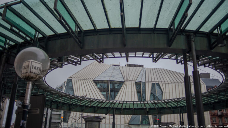 Circular glass canopy and taxi sign frame an angular modern building beneath radiating metal supports.