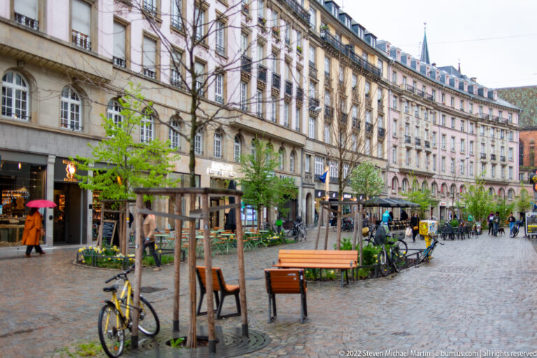 Cobbled Strasbourg pedestrian street with benches, young trees, parked bicycles, shops, and historic façades.