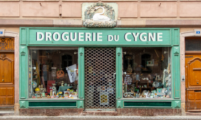 Turquoise Droguerie du Cygne storefront with swan emblem, display windows, and household goods.