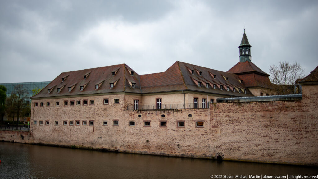 Long fortified masonry complex with small windows, steep tiled roofs, and a tower beside a river.