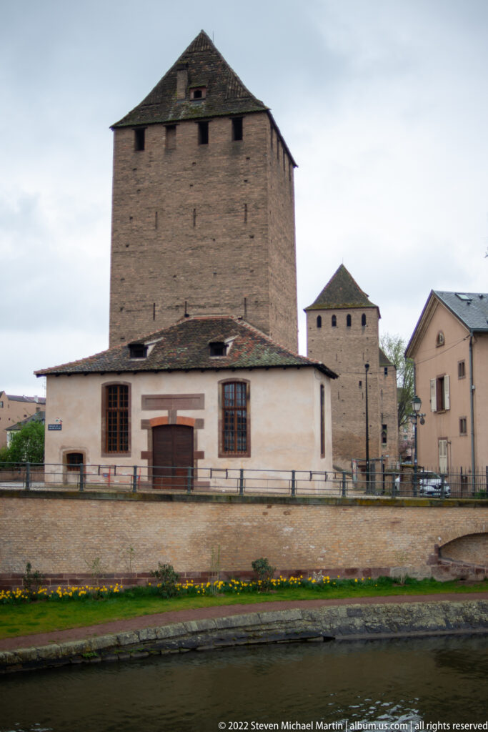 Two heavy masonry defensive towers with steep roofs stand beside a canal and low riverside buildings.
