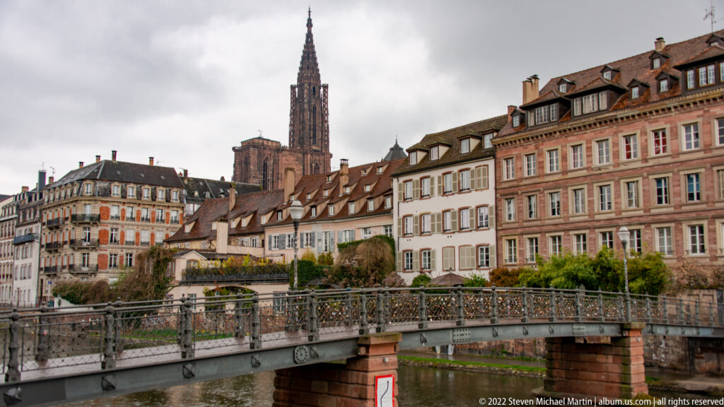 Pedestrian bridge over a Strasbourg canal with historic houses and the cathedral spire rising behind them.