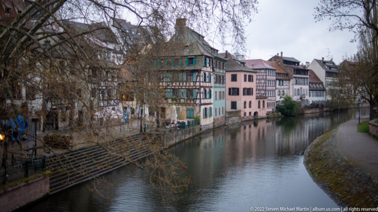 Half-timbered and pastel houses line a narrow Strasbourg canal with steps and bare trees along the quay.