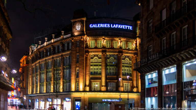 Illuminated Galeries Lafayette building at night with arched windows, corner clock, and blue rooftop sign.