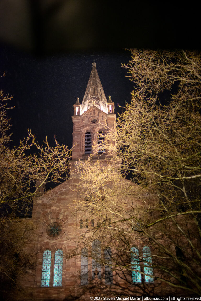 Illuminated historic church with square bell tower, arched windows, and bare tree branches at night.