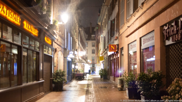 Narrow Strasbourg lane at night with glowing shop signs, warm streetlights, planters, and reflective paving.