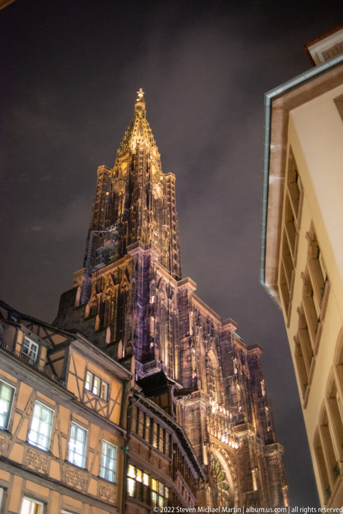 Illuminated Strasbourg Cathedral tower rises above nearby roofs and façades against the night sky.