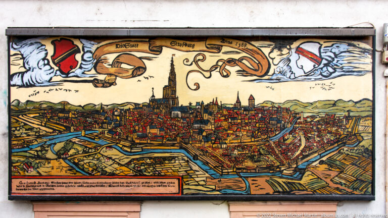 Illustrated historical bird’s-eye view of fortified Strasbourg with cathedral spire, walls, waterways, and heraldic emblems.