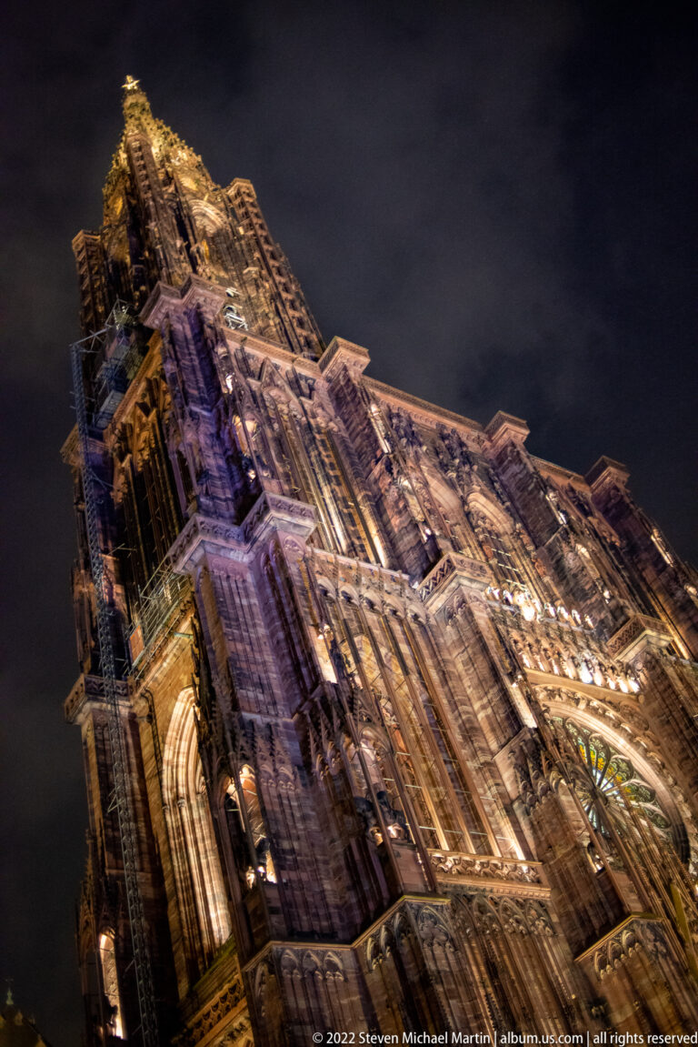 Steep upward nighttime view of Strasbourg Cathedral's illuminated Gothic tower against a dark sky.
