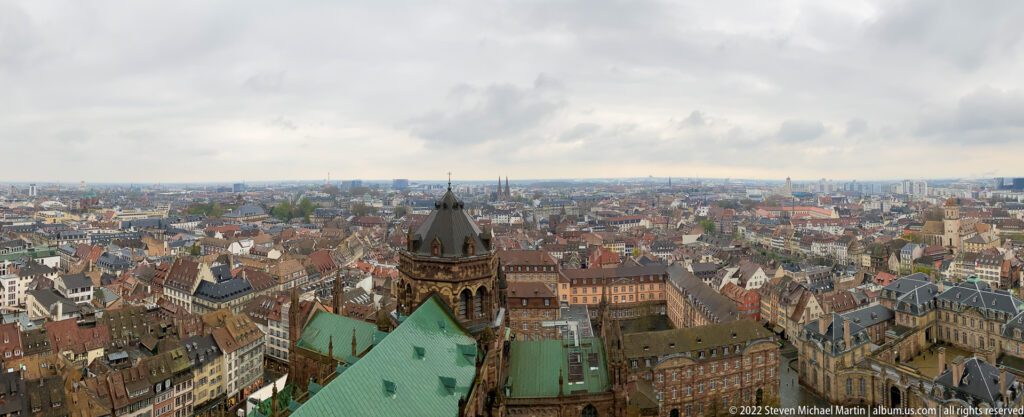Wide elevated panorama over Strasbourg rooftops with a nearby church tower and green roof in the foreground.