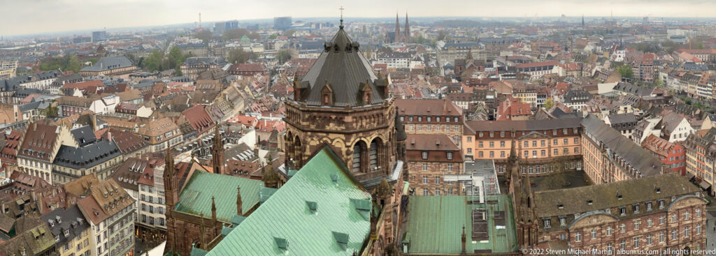 Elevated view over Strasbourg with a dark church tower, green copper roofs, and dense historic rooftops.