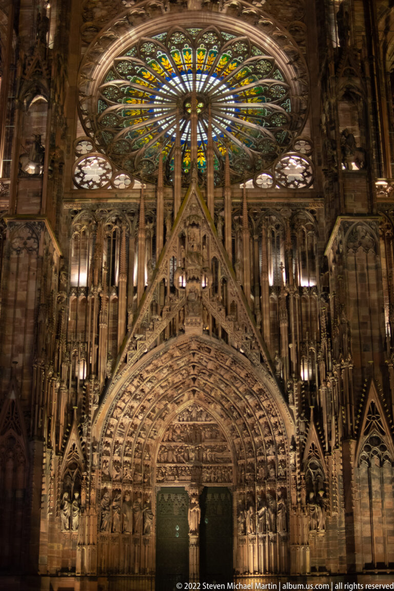 Illuminated Strasbourg Cathedral façade with sculpted central portal and large stained-glass rose window above.