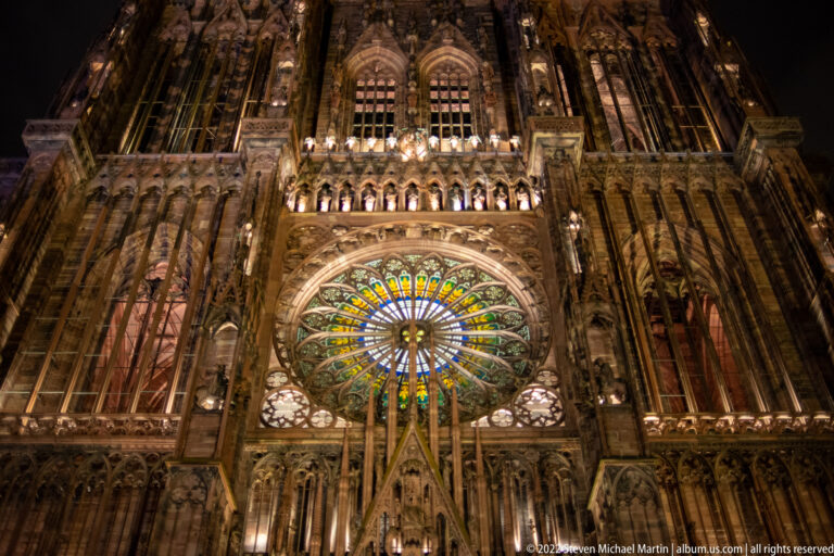 Large illuminated stained-glass rose window centered within the Gothic west façade of Strasbourg Cathedral.