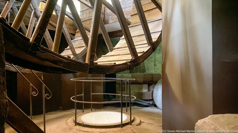 Large timber wheel structure above metal safety railings in a rough masonry upper interior of Strasbourg Cathedral.