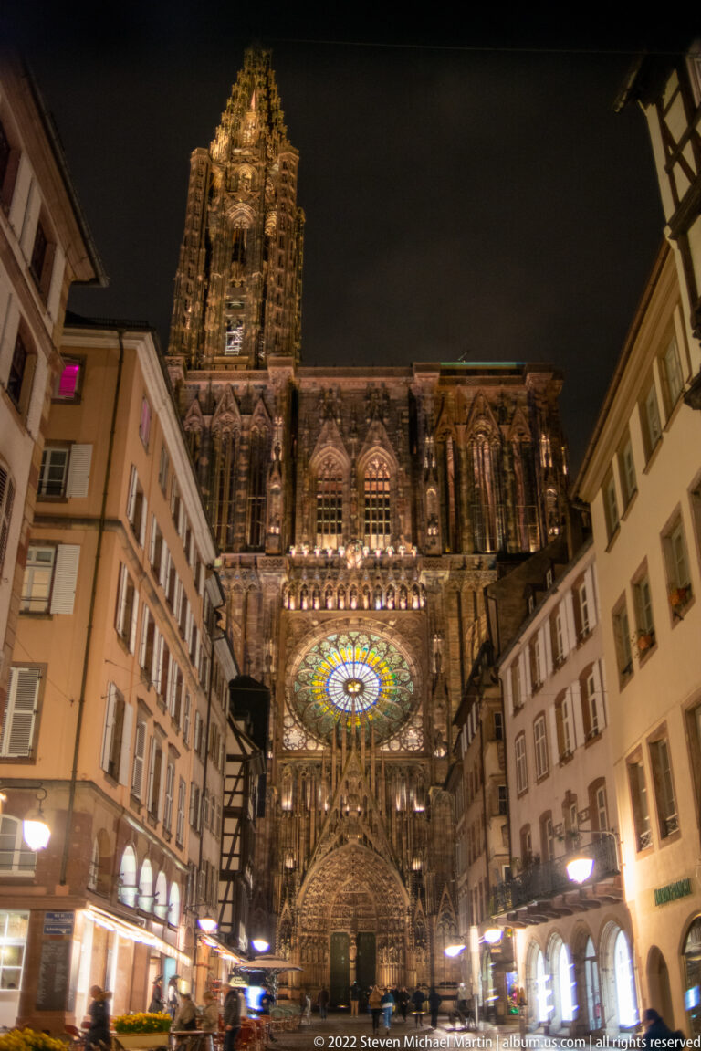 Strasbourg Cathedral illuminated at night, with glowing rose window and tower framed by neighboring buildings.