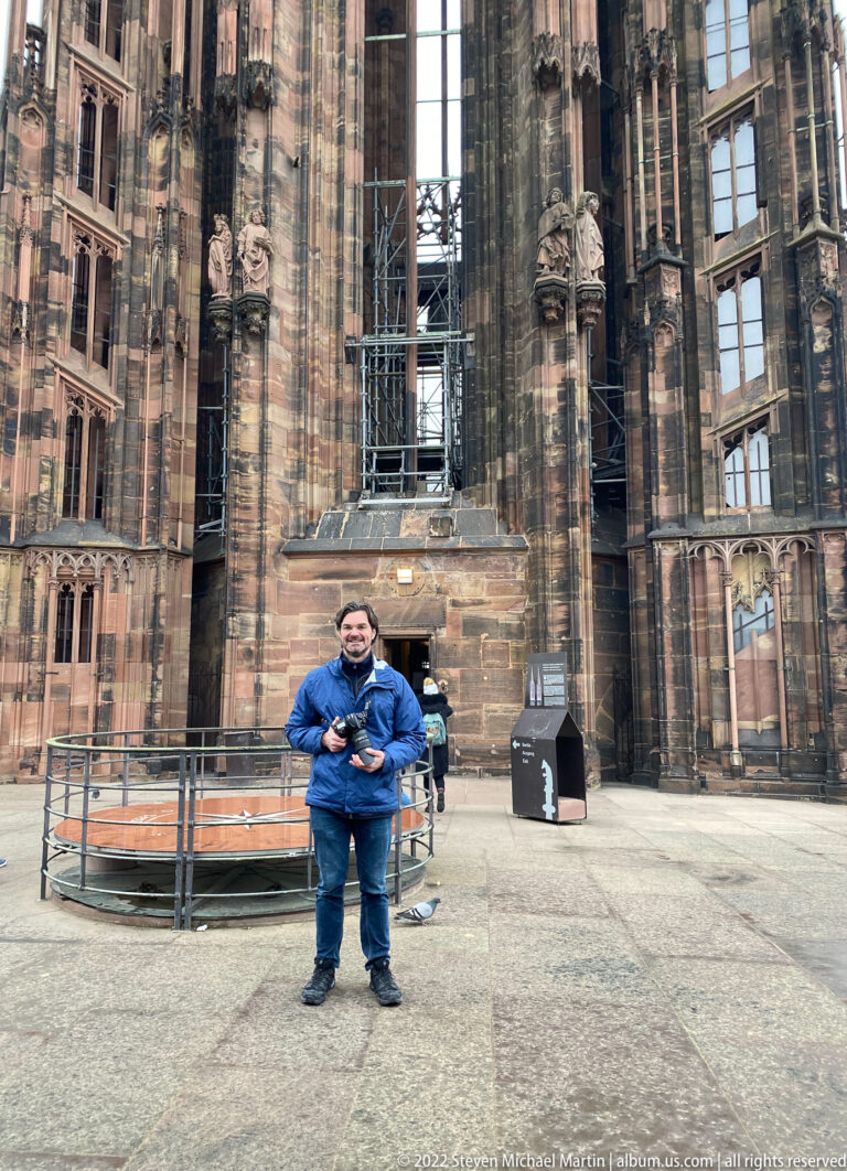 Visitor in a blue jacket stands on a stone platform amid the massive Gothic masonry of Strasbourg Cathedral.