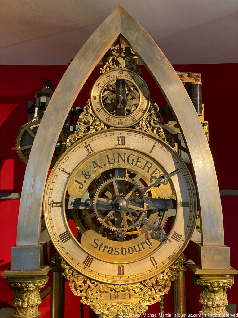 Exposed brass mechanical clock marked J. & A. Ungerer Strasbourg, with large dial, gears, and smaller upper dial.