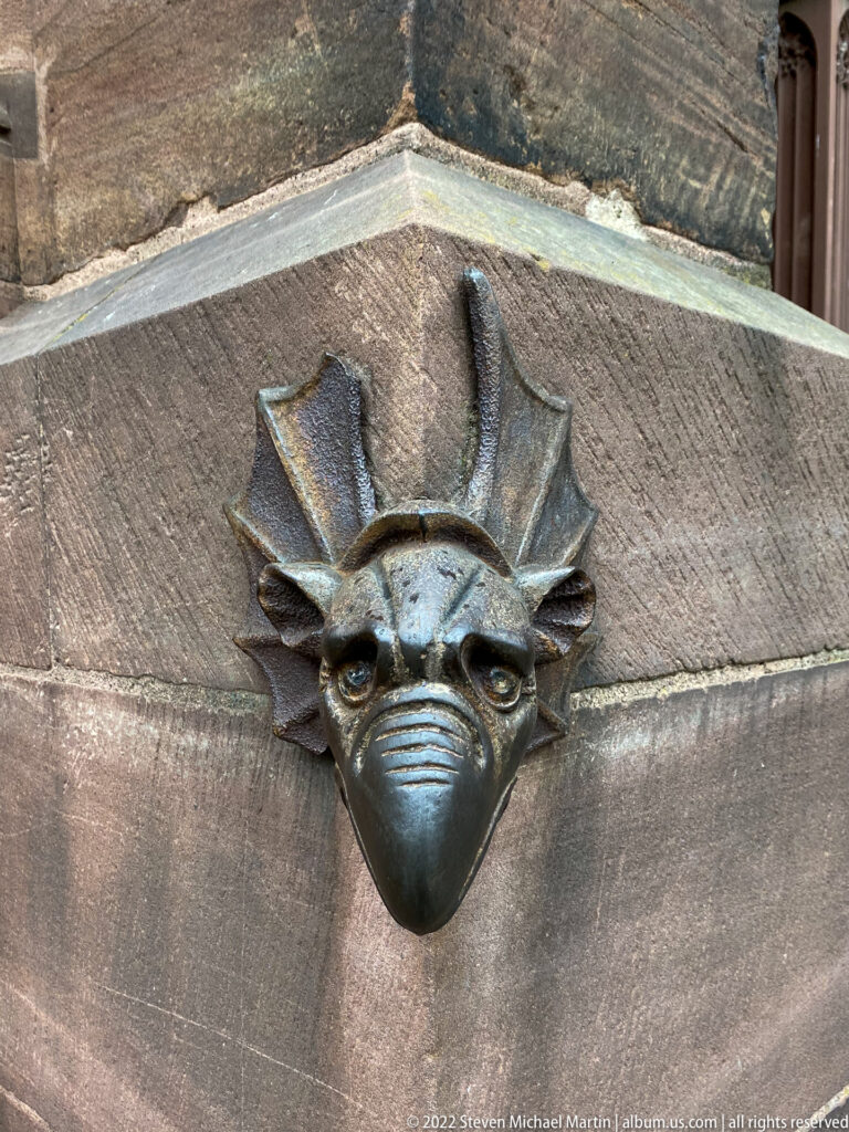 Dark metal grotesque with large eyes and pointed ears projecting from sloped pink sandstone.