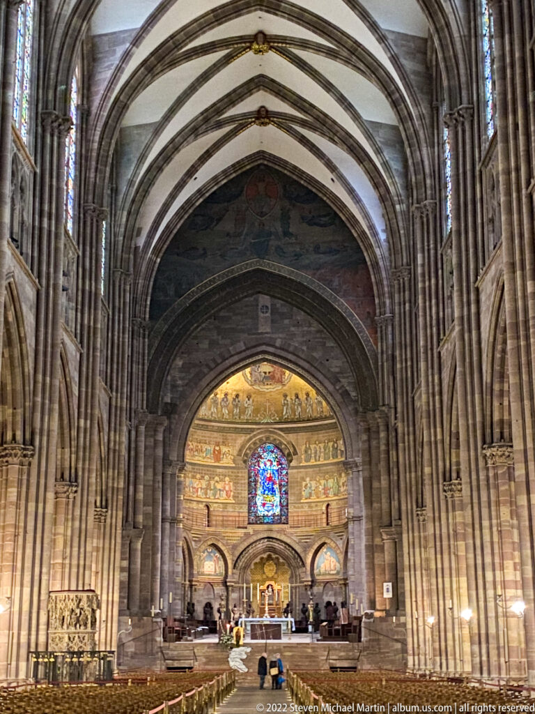 Stone-arched church nave leading toward a painted apse, stained-glass window, and raised sanctuary in Strasbourg.