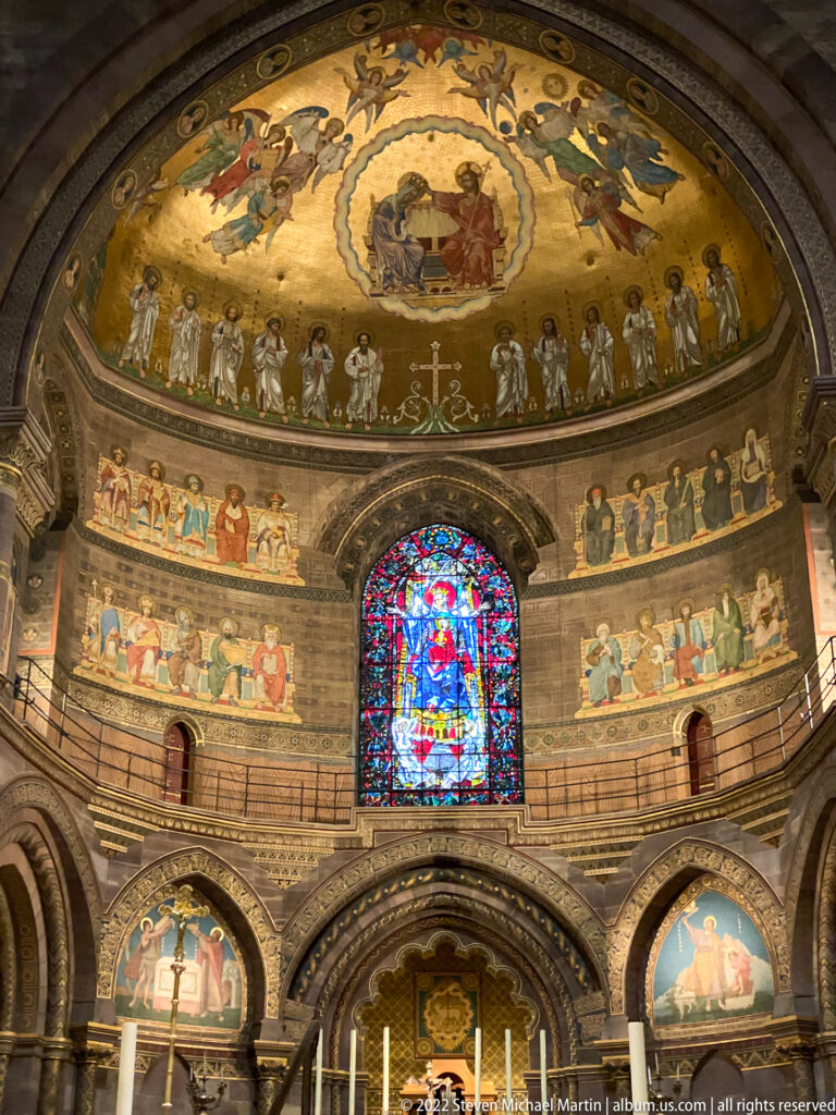 Painted church apse with Christ-centered semi-dome mural, bands of sacred figures, and a central stained-glass window.