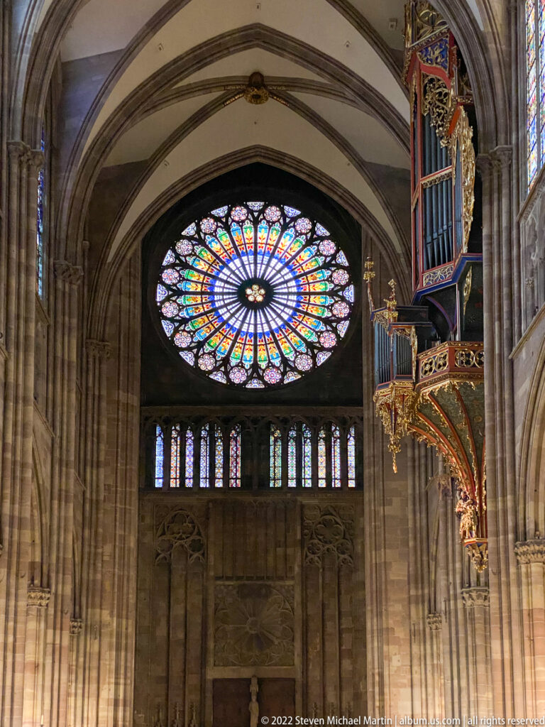 Large stained-glass rose window above lancets, with part of the colorful suspended organ case at right.