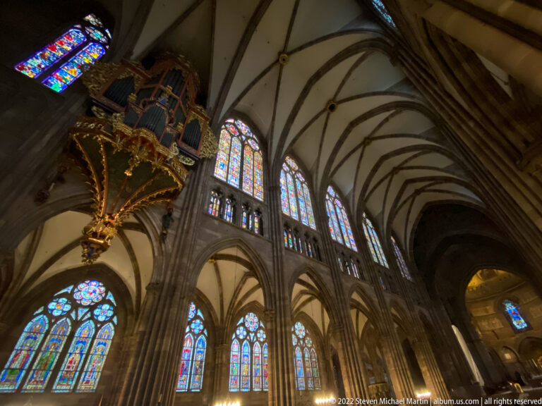 Low-angle cathedral interior with ribbed vaults, stained-glass windows, pointed arches, and suspended organ case.
