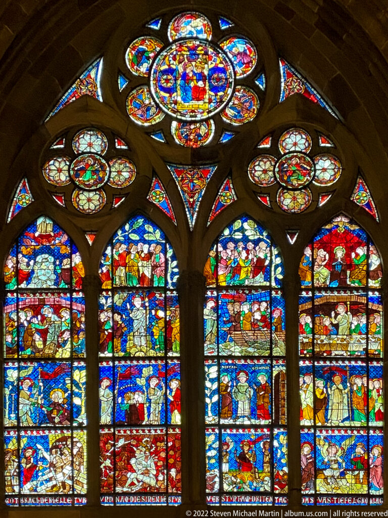 Four narrative stained-glass lancets beneath colorful circular quatrefoil windows in Strasbourg Cathedral.
