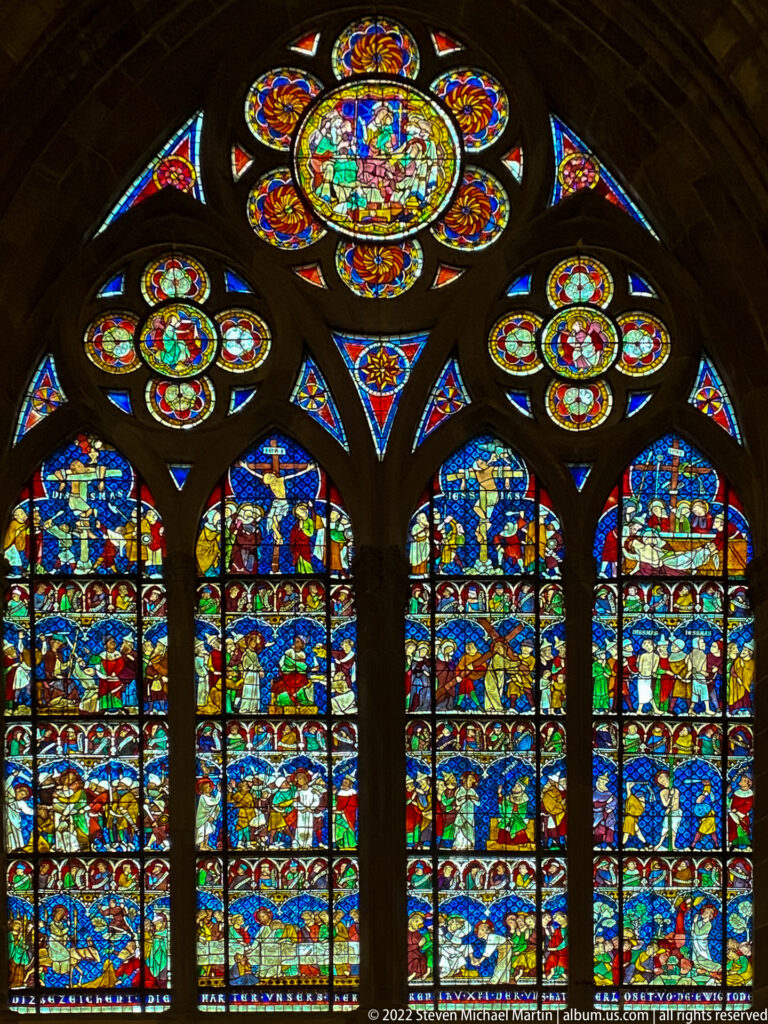 Four blue-dominant narrative stained-glass lancets beneath multifoil tracery windows in Strasbourg Cathedral.