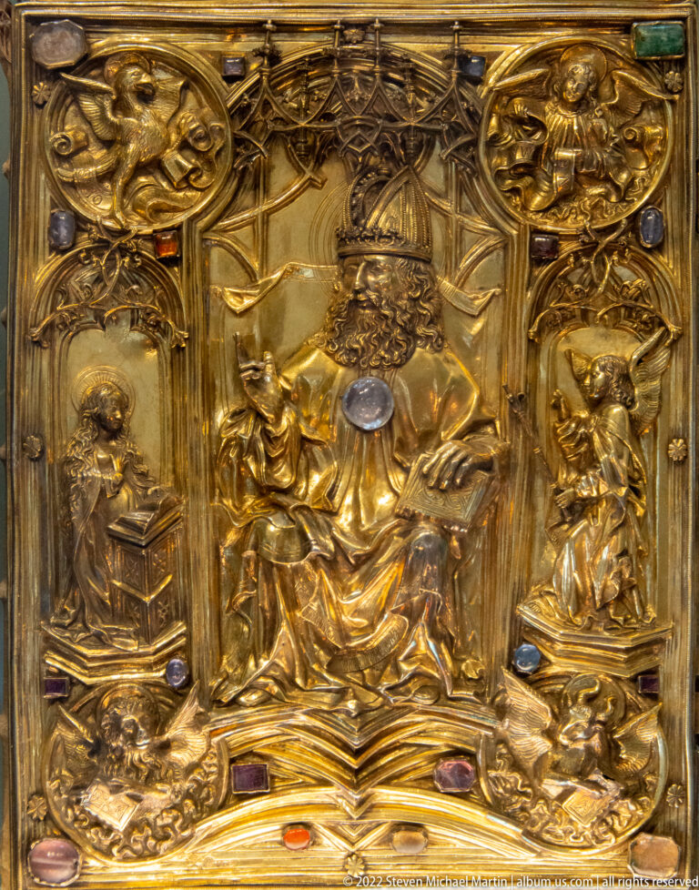 Close view of gilded relief metalwork with an enthroned central figure, smaller figures, and colored gemstones.